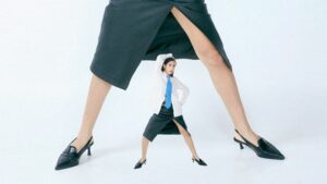 Tiny woman stands between giant legs in skirts.