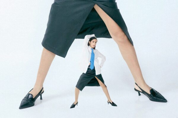 Tiny woman stands between giant legs in skirts.