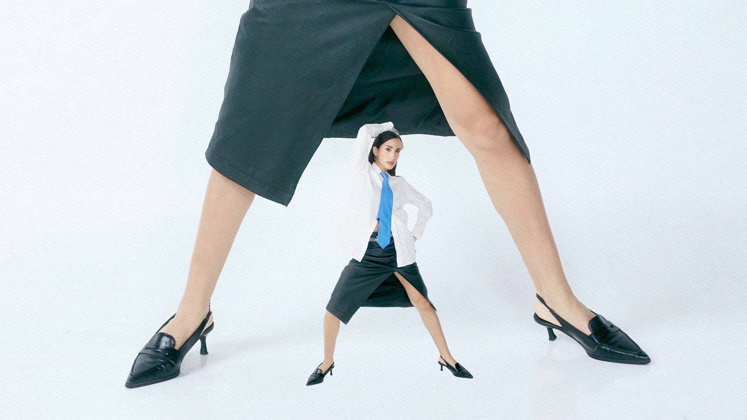 Tiny woman stands between giant legs in skirts.